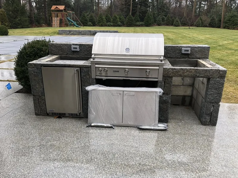 Granite-clad outdoor kitchen island with built-in grill and refrigerator being fitted on a Westchester patio