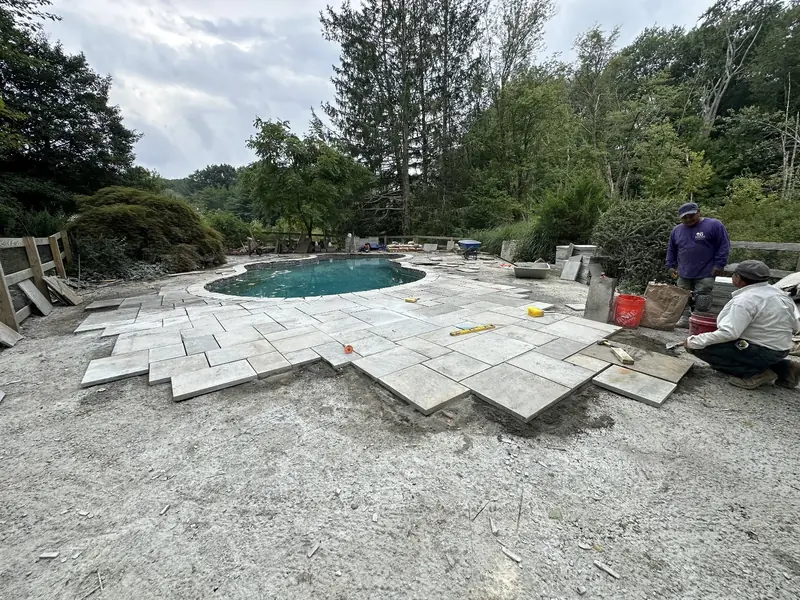 Two of the Baudi crew dry-laying large stone pavers on a bedding layer beside a pool