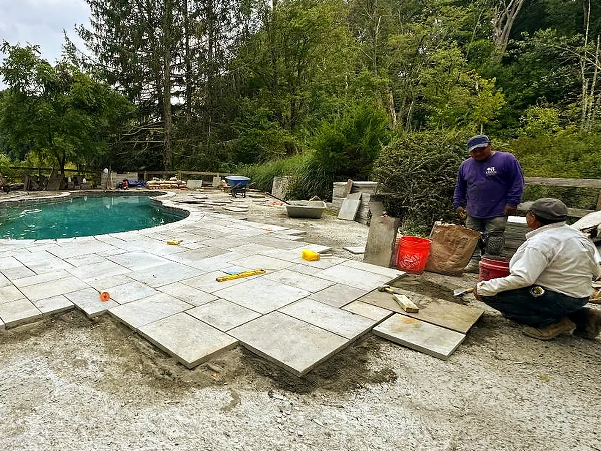 Crew setting large stone slabs on a bedding layer beside a filled pool
